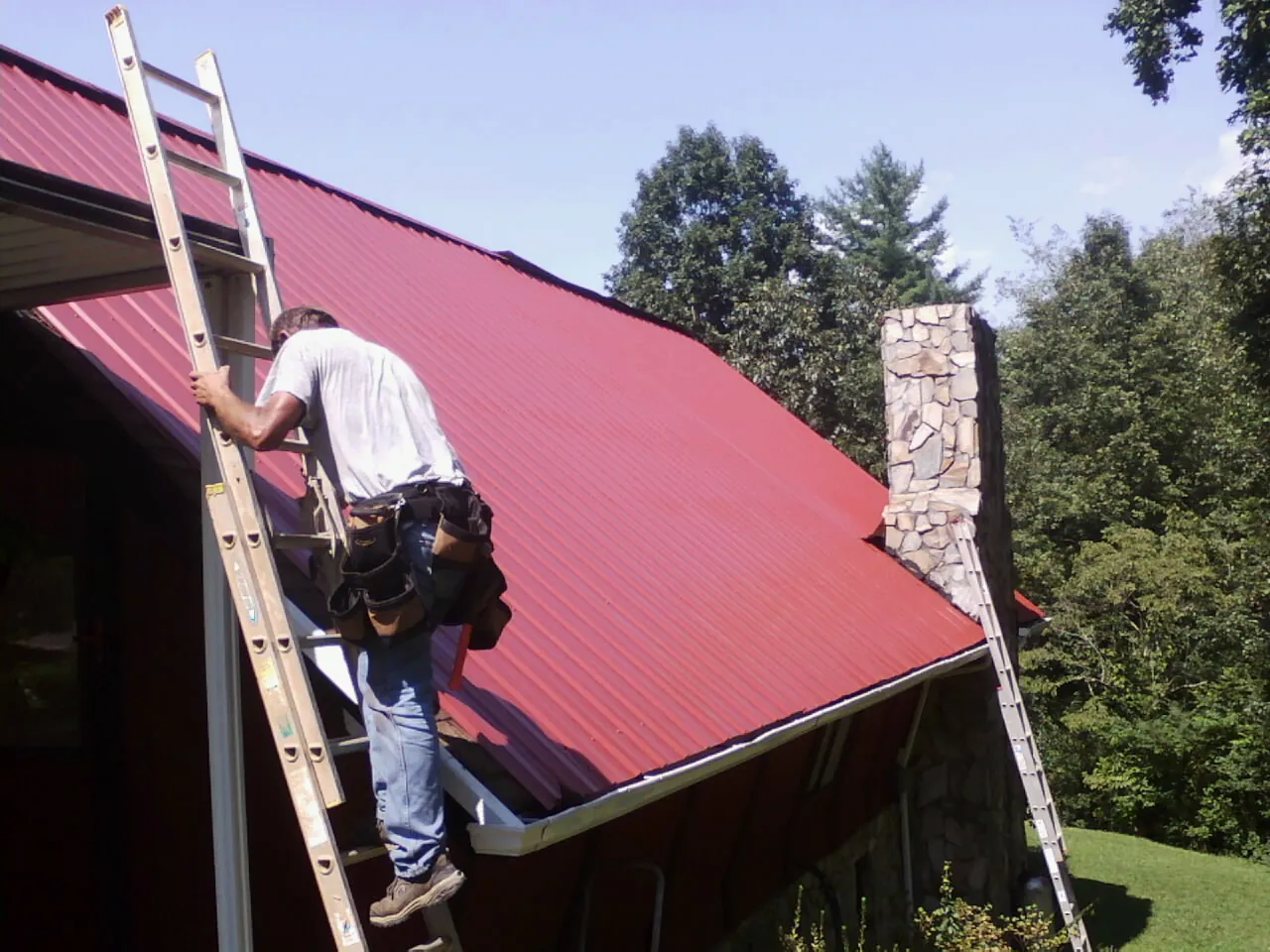 Roofer on a ladder beside a red metal roof and stone chimney — Soffit & Fascia Repair by Roofing Brothers in Autryville